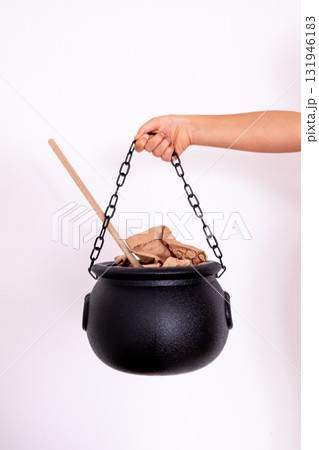 Hand holding black cauldron with wooden spoon and brown paper against white background Hand holding black cauldron with wooden spoon and brown paper against white background 131946183