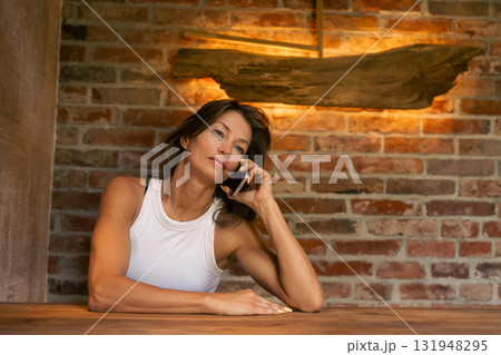 A reflective woman is engaged in a phone conversation while standing against a chic and stylish brick wall backdrop A reflective woman is engaged in a phone conversation while standing against a chic and stylish brick wall backdrop 131948295