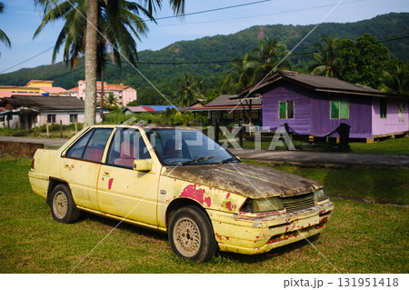 Old yellow car sits on grassy area with palm trees and colorful houses in background during sunny afternoon in rural setting 131951418