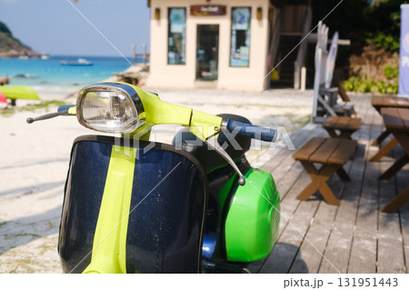 Bright scooter parked near a beachside cafe with clear blue water and wooden tables in the background during a sunny day 131951443