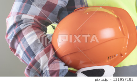 A person wearing a yellow and orange safety vest is holding an orange hard hat 131957193