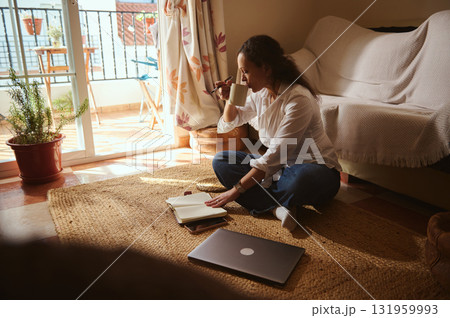 Woman Working From Home On Rug Sips Coffee, Writes In Notebook With Laptop Nearby 131959993