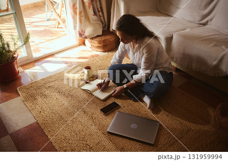 Woman Writing in Notebook at Home With Laptop, Coffee, and Sunlit Living Room 131959994