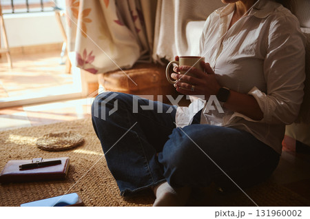 Relaxed Home Coffee Moment: Woman Sits on Floor With Mug in Sunlit Cozy Space Relaxed Home Coffee Moment: Woman Sits on Floor With Mug in Sunlit Cozy Space 131960002