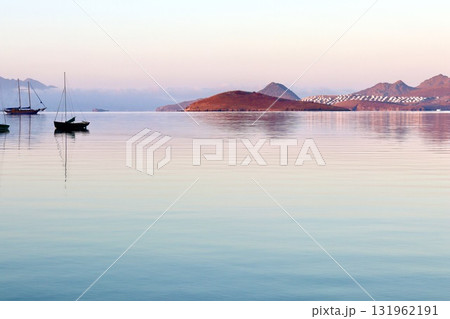 A peaceful sunrise or sunset over a calm, reflective sea, with sailboats and a small island in the distance, bathed in soft pastel light against a backdrop of mountains and a distant coastal town. A peaceful sunrise or sunset over a calm, reflective sea, with sailboats and a small island in the distance, bathed in soft pastel light against a backdrop of mountains and a distant coastal town. 131962191
