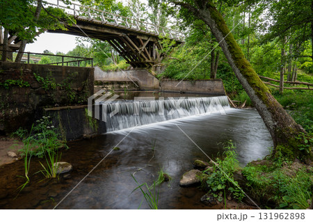 A serene and calm river gracefully flows beneath a rustic wooden bridge, enveloped by lush greenery and tranquil nature A serene and calm river gracefully flows beneath a rustic wooden bridge, enveloped by lush greenery and tranquil nature 131962898