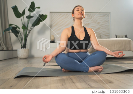 Yoga mindfulness meditation. Woman practicing yoga in living room at home. Plus size female sitting in lotus pose meditating smiling relaxing indoor. Girl doing breathing practice. Yoga at home 131963306