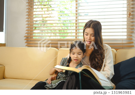 Happy Asian mother and little girl enjoying story time on the sofa, sharing smiles, love, and family togetherness 131963981