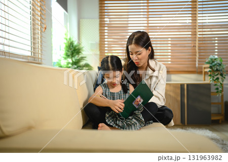 An Asian mother and daughter sit together indoors, smiling and bonding over a book 131963982