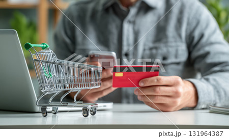 A close-up shot of a person making a digital payment for online shopping. They hold a red credit card and a smartphone near a laptop, with a miniature shopping cart in the foreground 131964387