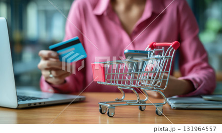 woman in pink shirt holding credit card and using laptop for online shopping. A miniature shopping cart holding a smartphone is placed prominently on the desk, symbolizing e-commerce, digital payment woman in pink shirt holding credit card and using laptop for online shopping. A miniature shopping cart holding a smartphone is placed prominently on the desk, symbolizing e-commerce, digital payment 131964557