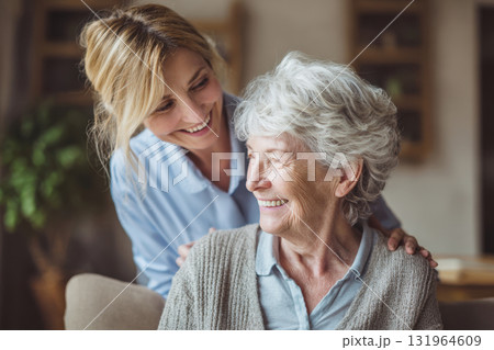 A warm, candid moment showing an elderly woman and a middle-aged woman sharing a smile and affectionate bond. The younger woman stands behind the senior, placing her hands on her shoulders A warm, candid moment showing an elderly woman and a middle-aged woman sharing a smile and affectionate bond. The younger woman stands behind the senior, placing her hands on her shoulders 131964609