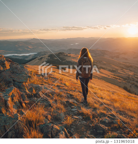 A female hiker with a backpack walks on a mountain ridge at sunset, with golden light illuminating the expansive landscape of rolling hills and distant lakes below 131965018