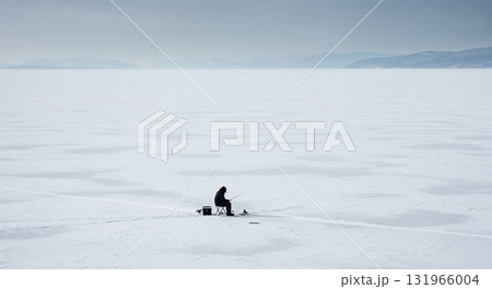 A lone person sits on a small chair, actively ice fishing on a vast, frozen, snow-covered lake under a grey, overcast sky with distant mountains. 131966004