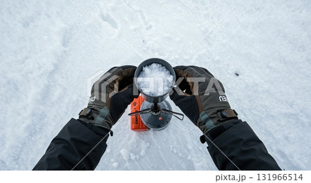 A person's dark gloved hands hold a silver pot full of white snow over a small orange camping stove, preparing water in a vast snowy outdoor environment. A person's dark gloved hands hold a silver pot full of white snow over a small orange camping stove, preparing water in a vast snowy outdoor environment. 131966514