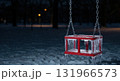 A red children's swing seat hangs heavily covered in glistening icicles and frost in a snow-covered park at night, illuminated by distant warm streetlights. 131966573