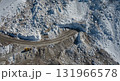 A yellow bulldozer navigates a winding dirt road cut through a rugged, snow-covered mountain landscape under bright sunlight, highlighting industrial work in extreme conditions. 131966578