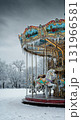 A classic ornate carousel featuring white horses and warm lights stands covered in snow and icicles within a serene winter park under an overcast sky. 131966581