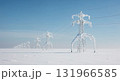 A long series of towering metal electricity pylons, heavily encrusted with white snow and ice, extends across a vast, pristine, flat winter landscape under a bright blue sky. 131966585