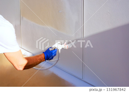 Handyman is applying fresh white paint to wall using spray gun in well lit room. Handyman is applying fresh white paint to wall using spray gun in well lit room. 131967842