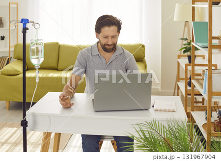 Young man working on a laptop at the desk at home or in office while receiving IV vitamin therapy. Young man working on a laptop at the desk at home or in office while receiving IV vitamin therapy. 131967904