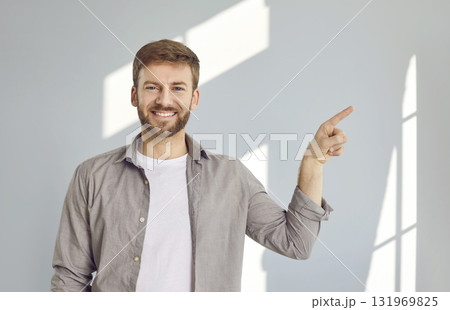 Young man pointing to copy space and looking at camera with smiling face on gray wall background. 131969825