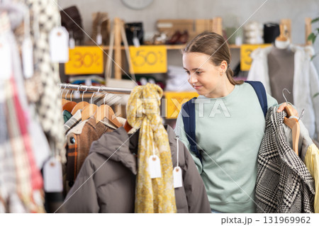 Positive young girl standing in interior of clothing boutique and choosing something on Black Friday sale of winter clothing collection 131969962