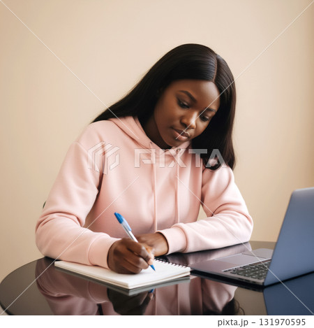 african american woman in a pink hoodie writes in a notebook at a table with a laptop 131970595