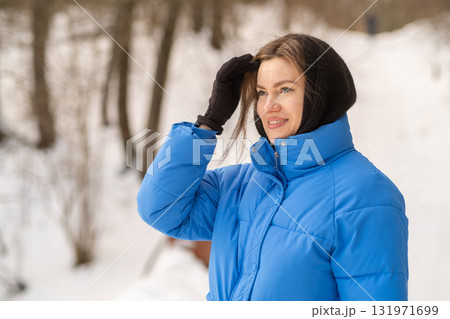 A young woman wearing a blue coat and gloves stands in a snowy landscape, smiling while brushing hair from her forehead under winter sunlight. 131971699