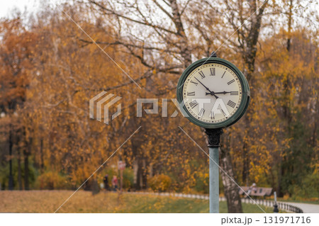 A vintage clock stands tall in a park, framed by vibrant autumn leaves as people stroll by in the evening light. 131971716