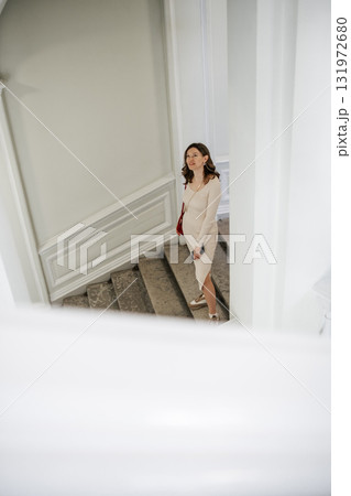 A fashionable woman in a beige dress stands on the staircase, elegantly looking upwards in a well-lit interior with neutral decor and classic architecture. A fashionable woman in a beige dress stands on the staircase, elegantly looking upwards in a well-lit interior with neutral decor and classic architecture. 131972680