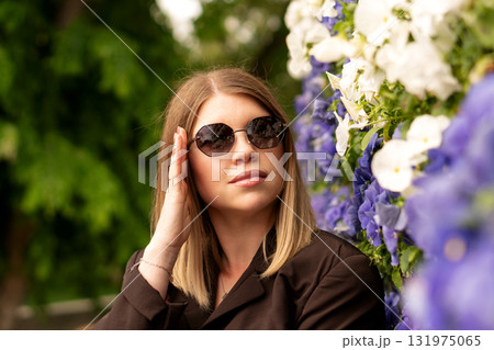 Stylish young woman adjusts sunglasses outdoors by floral backdrop. Stylish young woman adjusts sunglasses outdoors by floral backdrop. 131975065