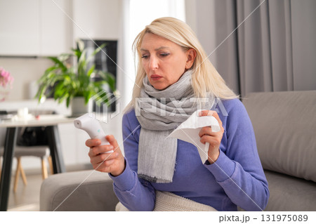 Blonde middle aged woman lying on a sofa feeling unwell blowing her nose with tissue and checking her temperature with a thermometer while staying at home Blonde middle aged woman lying on a sofa feeling unwell blowing her nose with tissue and checking her temperature with a thermometer while staying at home 131975089