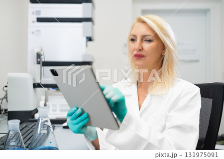 Blonde woman without mask wearing gloves recording laboratory experiment results on a tablet while standing in a chemistry lab with flasks, beakers, and HPLC equipment 131975091
