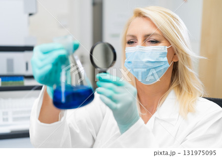 Middle aged female scientist wearing mask in lab, studying chemical solution in flask through magnifying glass. Middle aged female scientist wearing mask in lab, studying chemical solution in flask through magnifying glass. 131975095