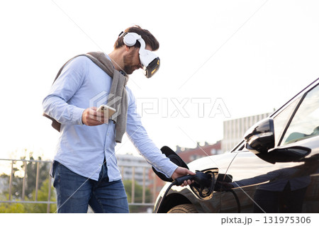 A man wearing VR glasses connects an electric vehicle to a charging station while holding a phone. 131975306