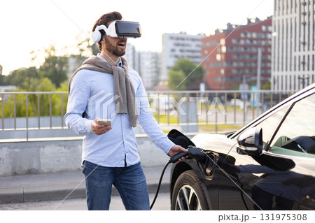 A man wearing a VR headset connects an electric vehicle to a charging station, holding his phone. A man wearing a VR headset connects an electric vehicle to a charging station, holding his phone. 131975308