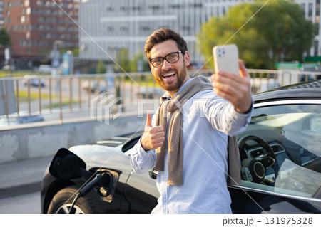 A man smiles while taking a selfie next to his electric vehicle while it's charging at a charging station. 131975328