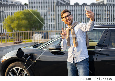 A man smiles and makes a peace sign while taking a selfie next to his electric vehicle as it charges at a charging station. A man smiles and makes a peace sign while taking a selfie next to his electric vehicle as it charges at a charging station. 131975330