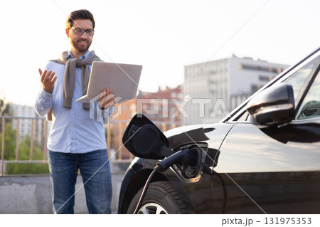 A man stands next to his electric vehicle while it charges, holding a laptop and appearing to be working or video conferencing. A man stands next to his electric vehicle while it charges, holding a laptop and appearing to be working or video conferencing. 131975353
