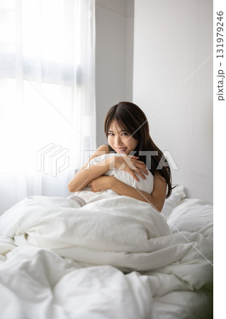 朝日を浴びながら、ベッドで枕を抱いて微笑む若い女性 朝日を浴びながら、ベッドで枕を抱いて微笑む若い女性 131979246