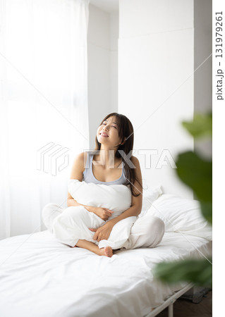 朝日を浴びながら、ベッドで枕を抱いて微笑む若い女性 朝日を浴びながら、ベッドで枕を抱いて微笑む若い女性 131979261