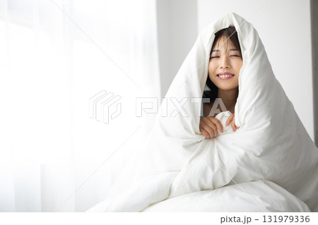 朝日を浴びながら、布団にくるまって微笑む若い女性 朝日を浴びながら、布団にくるまって微笑む若い女性 131979336