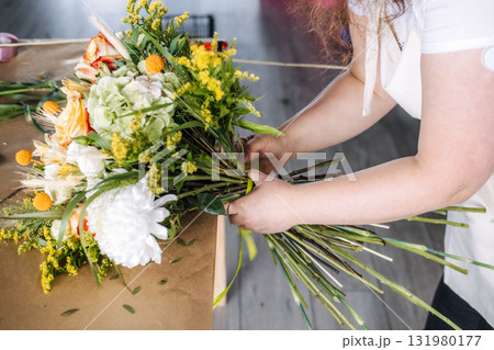 Florist tying ribbon around stems of a fall-colored bouquet on a wooden table. Modern bouquet design, autumn color palette, botanical craft, seasonal floral artistry 131980177