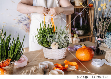 Woman arranging white heather and evergreen branches into a decorative basket on a craft table. Thanksgiving workshop, seasonal DIY, natural decor, creative wellness 131980178