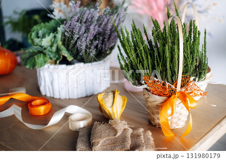 Small woven baskets with evergreen sprigs and floral arrangements sit on a kraft paper-covered table with ribbons and gourds. Thanksgiving flower workshop, DIY fall bouquet, florist workspace 131980179
