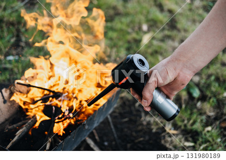 Hand igniting a wood fire in a metal grill setup using a portable air blower outdoors. Fall grilling season, backyard BBQ, outdoor chef lifestyle, fire pit recipes 131980189