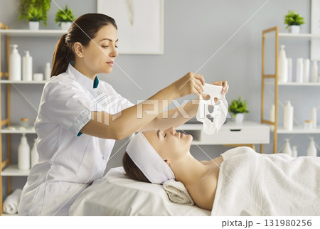 Female beautician applying moisturizing sheet mask on young woman face in beauty spa salon. 131980256