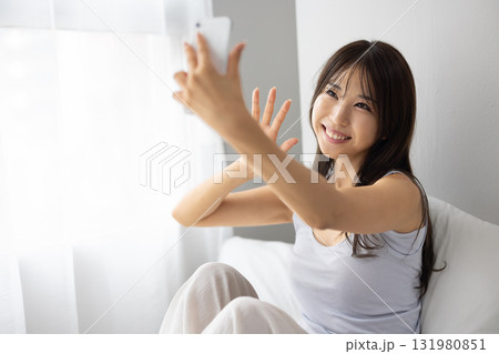 ベッドの上で窓からの朝日を浴びながらスマートフォンでビデオ通話する若い女性若い女性 ベッドの上で窓からの朝日を浴びながらスマートフォンでビデオ通話する若い女性若い女性 131980851