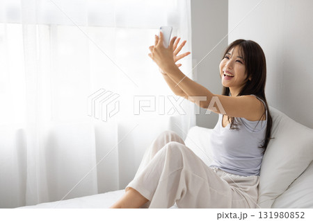 ベッドの上で窓からの朝日を浴びながらスマートフォンでビデオ通話する若い女性若い女性 ベッドの上で窓からの朝日を浴びながらスマートフォンでビデオ通話する若い女性若い女性 131980852